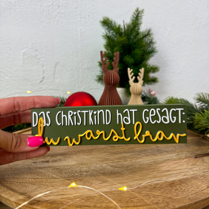 Christkind-Spruch-Schild in Weiß und Gold in einer Hand gehalten als Größenvergleich vor Weihnachtsdeko