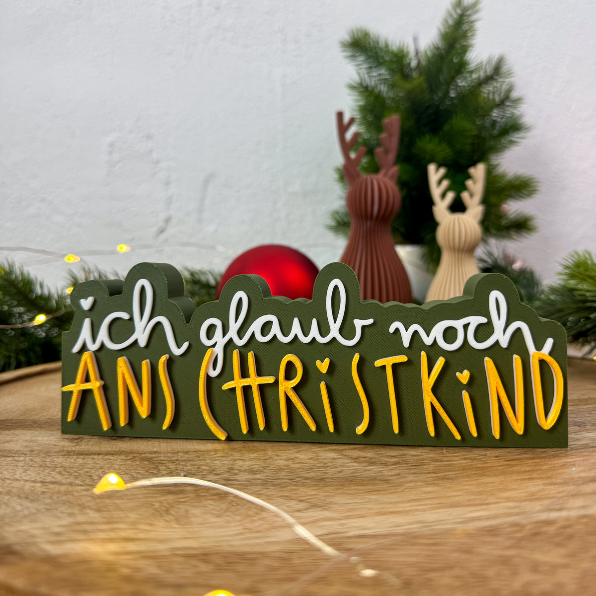 Christkind-Spruch-Schild „ich glaub noch ans Christkind" mit Rentierfiguren im Hintergrund