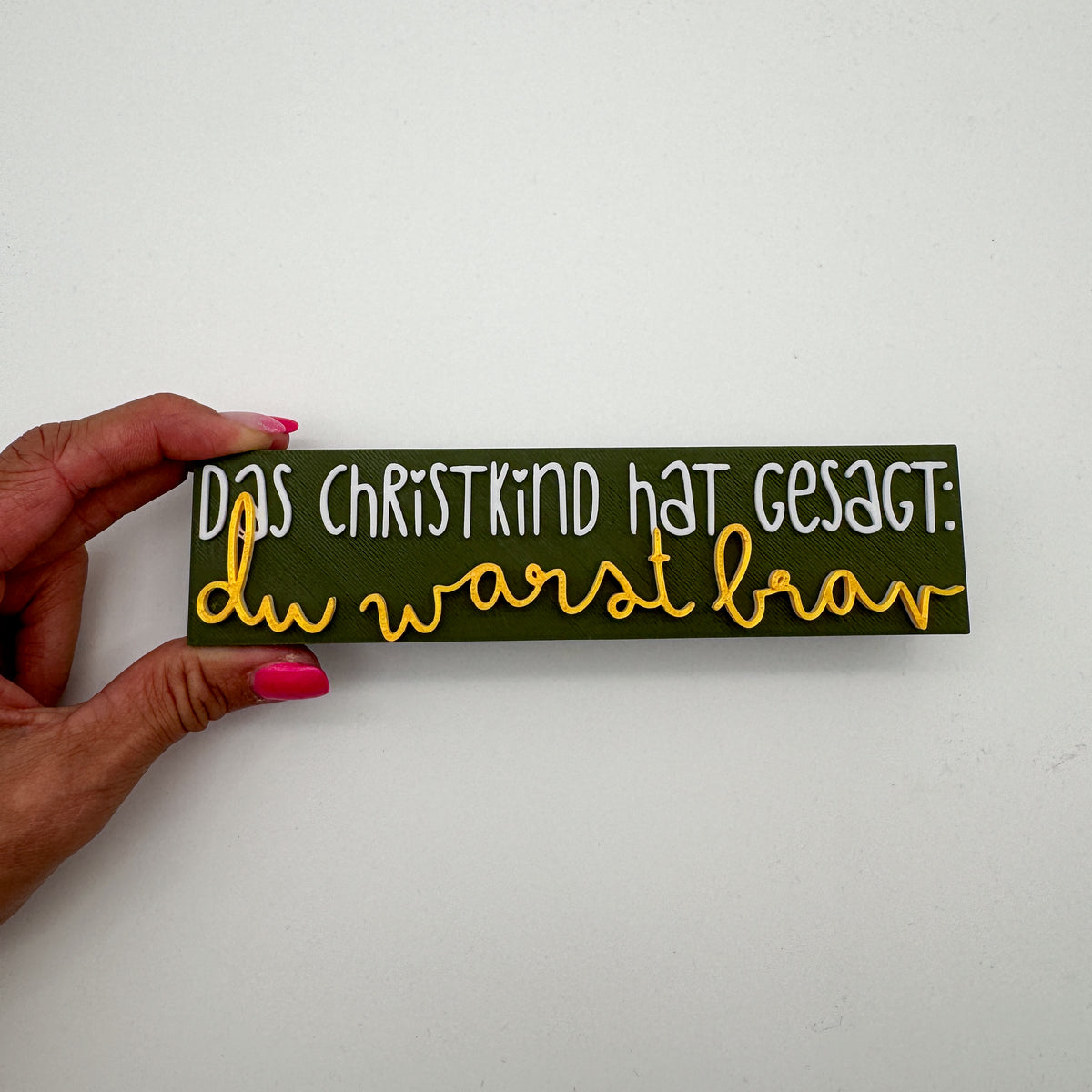 Nahaufnahme vom Christkind-Spruch-Schild in Olivgrün mit weißer und goldener Schrift, in der Hand gehalten