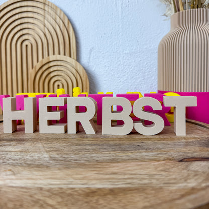 3D Buchstaben HERBST in Beige freistehend auf Holz, dahinter dieselben Deko-Buchstaben in Pink und Gelb