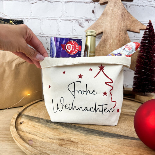 Hand hält einen Geschenkkorb „Frohe Weihnachten“, gefüllt mit Tee und einer Sektflasche. Ein ideales kleines Weihnachtsgeschenk für die Kollegin.