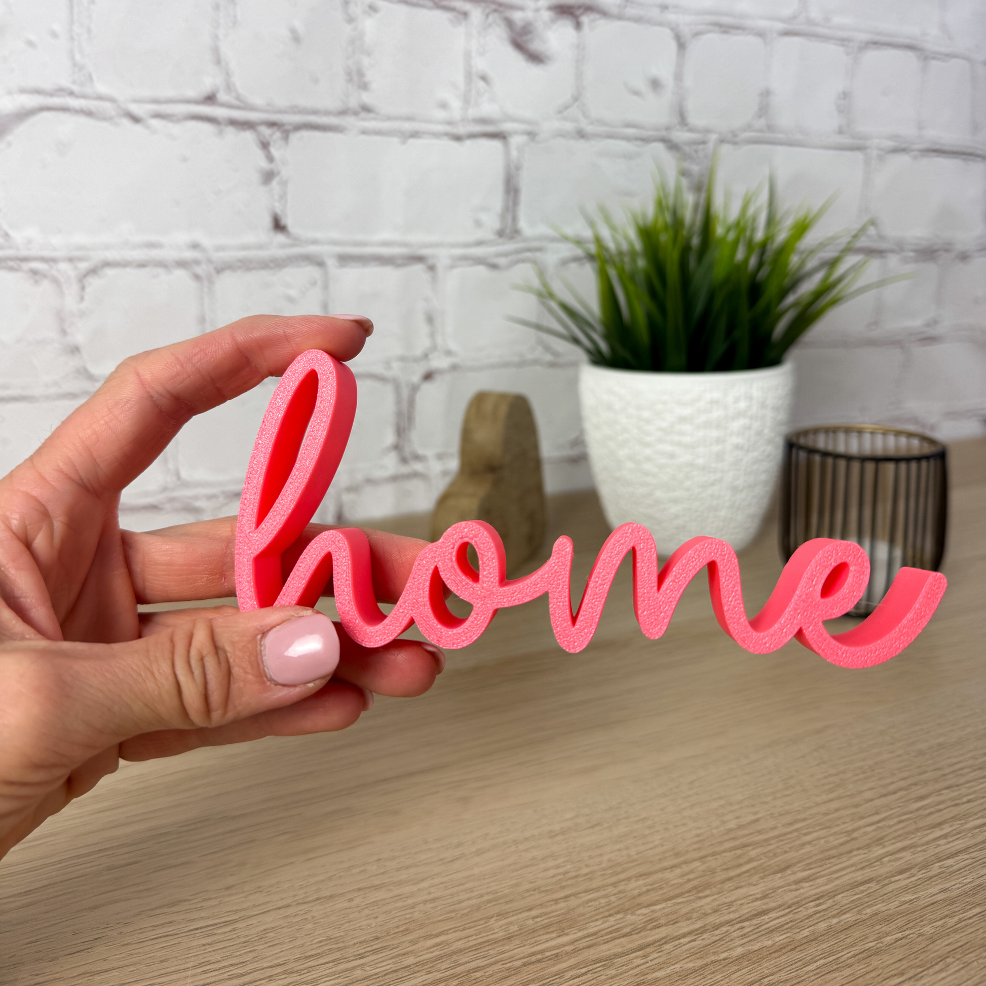 Pinkfarbener 3D-Schriftzug 'home' wird von Hand gehalten, im Hintergrund Pflanzen-Deko – handgemachte Dekoidee für Zuhause.