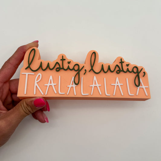 Schild „lustig lustig tralalala" in Apricot, Dunkelgrün und Weiß in einer Hand gehalten