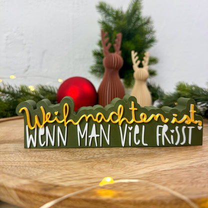 Lustiger Weihnachtsspruch „Weihnachten ist, wenn man viel frisst" in Nahaufnahme auf Holztisch