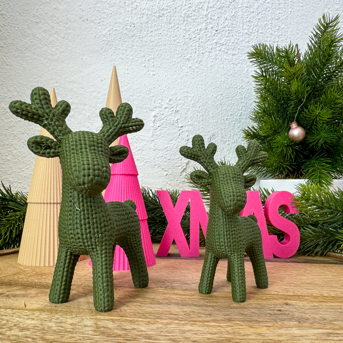 Zwei Rentier Figuren in Strickoptik neben Deko-Bäumen und pinkem „XMAS"-Schriftzug