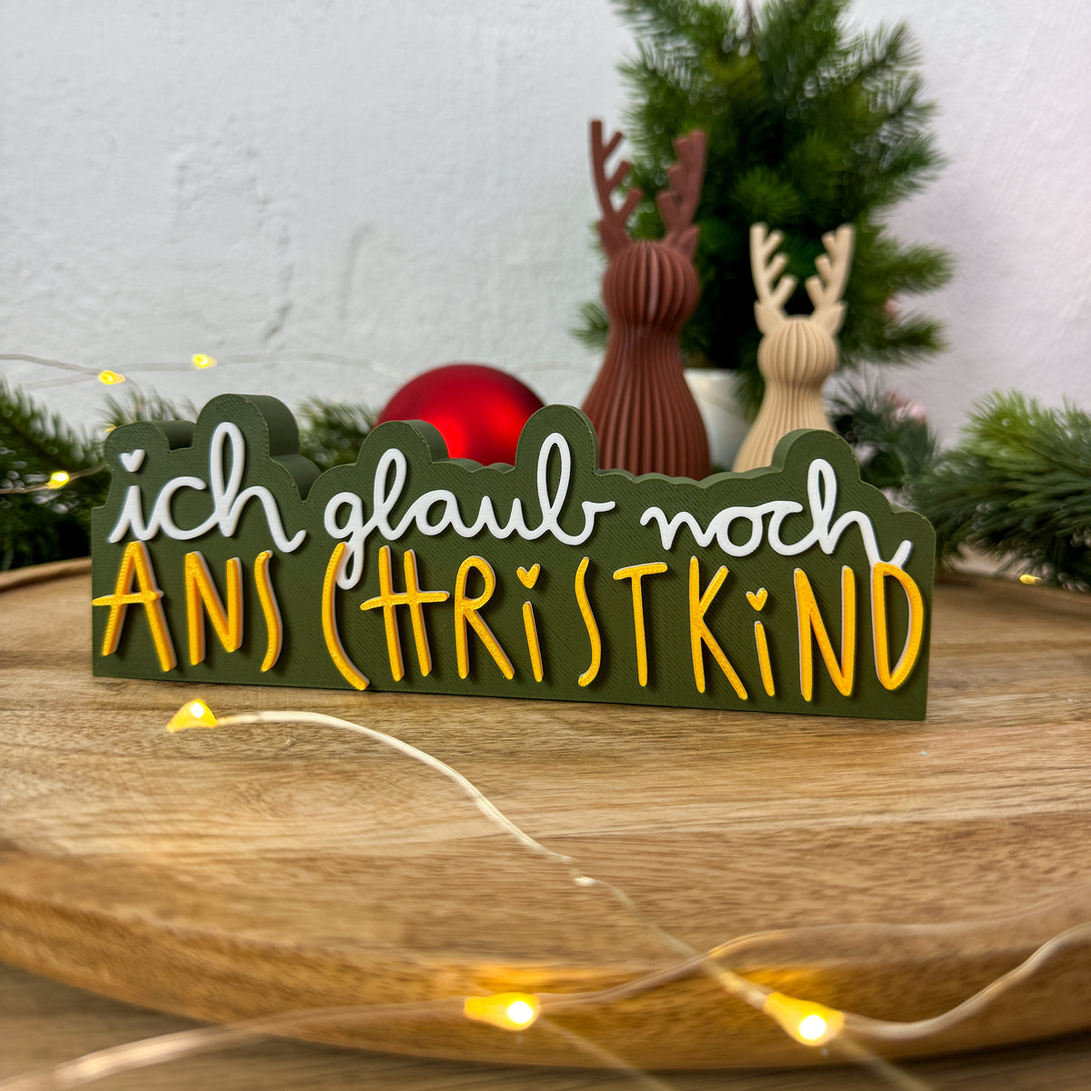 Weihnachtsdeko-Spruch „ich glaub noch ans Christkind" in Weiß und Gold auf dunkelgrünem Riegel
