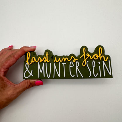 Weihnachtsdeko-Spruch-Schild in Dunkelgrün, Gold und Weiß in einer Hand gehalten