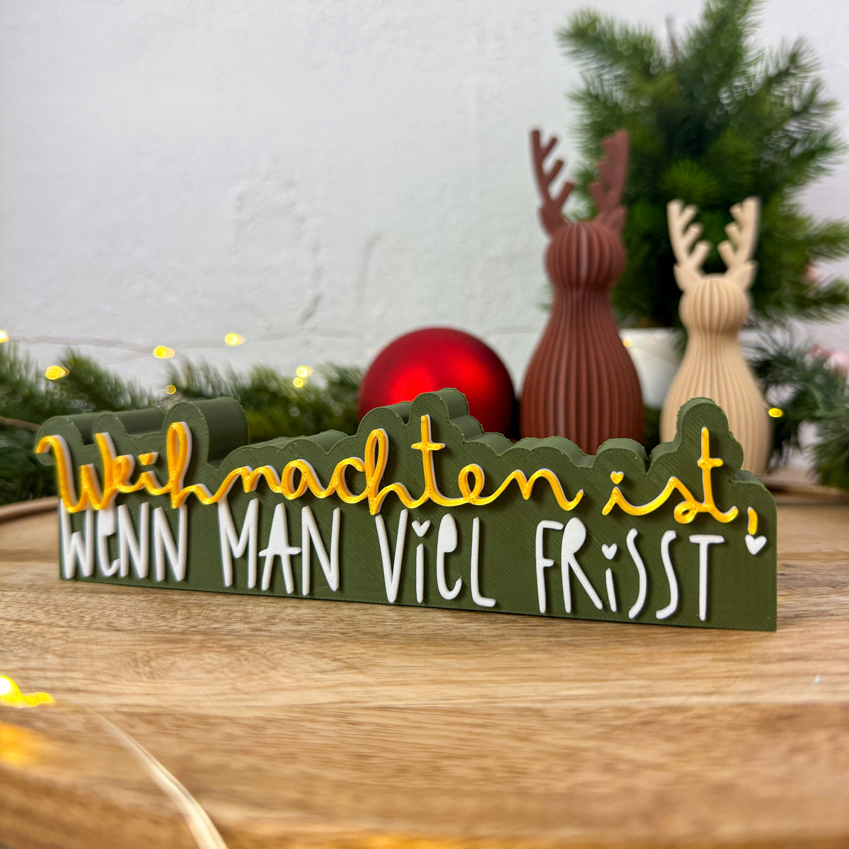 Weihnachtsspruch „Weihnachten ist, wenn man viel frisst" in Dunkelgrün, Gelb und Weiß neben Weihnachtsbaum und Rentierfiguren